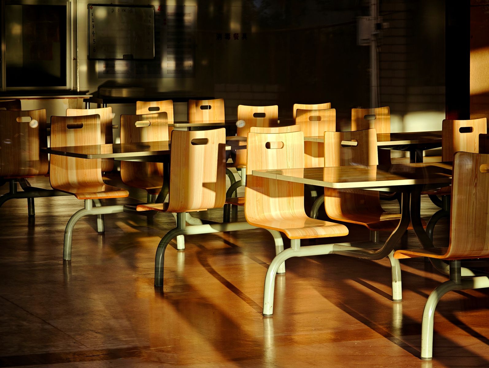 Empty wooden chairs and tables in a sunlit cafeteria setting, casting dramatic shadows.